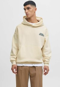 Cream hoodie with a large pocket, ribbed cuffs, and a logo in dark green on the chest. Worn over a white shirt and paired with brown pants.