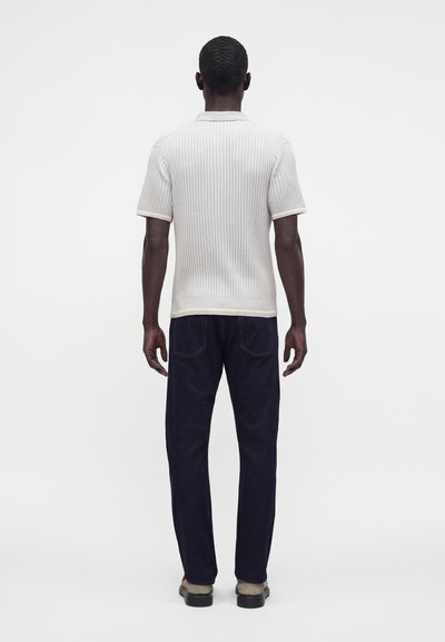 Homme debout de dos portant un pull en maille blanc à manches courtes, un pantalon bleu foncé et des chaussures claires sur un fond uni.