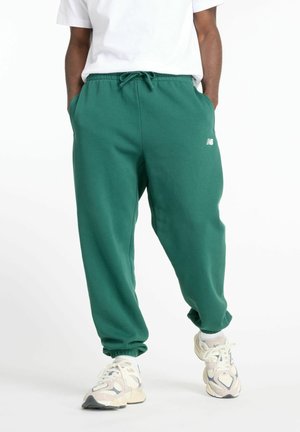 Mann trägt grüne Jogginghosen mit elastischen Bündchen, weißes T-Shirt und weiße Sneakers, steht mit den Händen in den Taschen vor weißem Hintergrund.