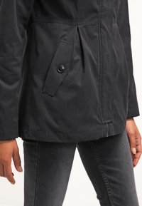 Person som bär en svart jacka med knäppt ficklock och blixtlås, tillsammans med mörkgråa slim fit-jeans, med händerna avslappnat vid sidorna.