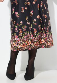 Blumenkleid mit schwarzem Hintergrund, verziert mit bunten Blumen, fließendem Design und knöchellangem Saum. Kombiniert mit schwarzen High Heels.