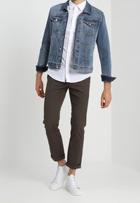 Jeansjacka i ljusblått med knapplås framtill och två bröstfickor, kombinerad med en vit skjorta, bruna byxor och vita sneakers.