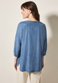 Top in denim azzurro chiaro con maniche 3/4, scollatura rotonda e fessure laterali. Texture liscia con una finitura uniforme.