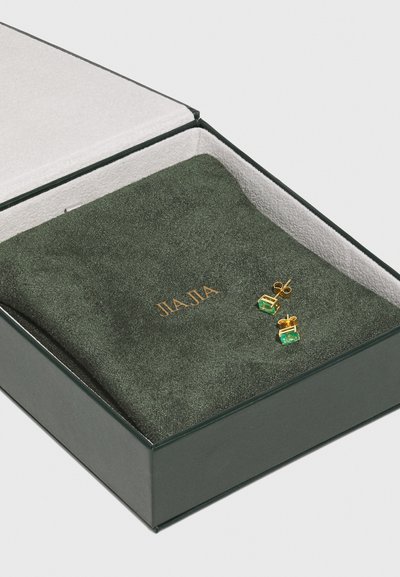 Vihreä samettinen korurasia, joka sisältää kultaisia nappikorvareita, joissa on emerald-leikkauksella leikattuja vihreitä kiviä. Rasiassa on kultalogo ja valkoinen sisus.
