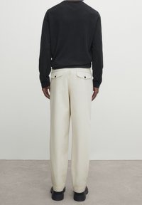 Personne portant un pantalon beige à jambes larges avec des poches arrière boutonnées, un haut à manches longues noir et des chaussures noires, se tenant sur un sol en béton.