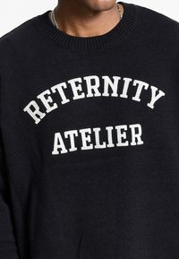 Sort strikket sweater med teksten "RETERNITY ATELIER" i hvid, med rund halsudskæring og struktureret stof.