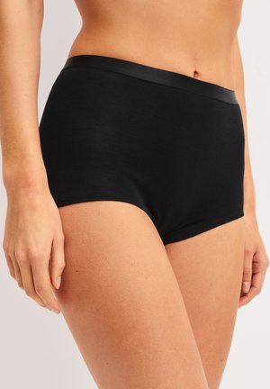 Femme portant des sous-vêtements noirs sans couture à taille haute avec une ceinture lisse, debout devant un fond uni.