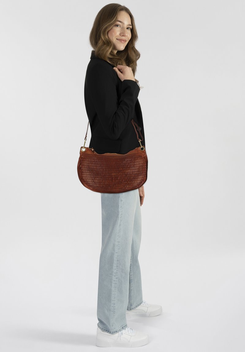 Geplottete braune Leder-Schultertasche mit geschwungener Form und goldenen Beschlägen, kombiniert mit hellblauen Jeans und weißen Sneakers.
