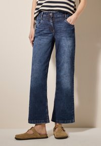 Wijdere blauwe denim jeans met een lichte vervaging, voorzakken en een knoopsluiting. Gecombineerd met tan open-toed schoenen.