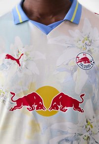 Maillot domicile du FC Salzbourg avec un design floral bleu clair, un logo Puma rouge et un graphique de taureau rouge sur fond de cercle jaune.