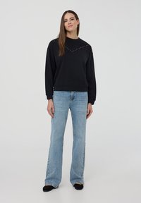 Felpa nera con scollo rotondo, decorata con piccoli chiodini disposti in un motivo a zig-zag, abbinata a jeans flare azzurri chiari.