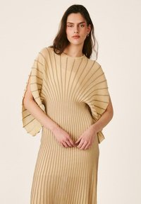 Mujer joven con el pelo largo y oscuro que lleva un vestido beige de punto acanalado con mangas cortas anchas y acampanadas, situada frente a un fondo neutro.