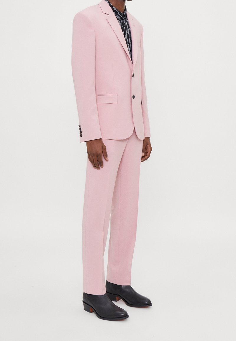 Costume rose clair avec coupe ajustée, blazer à un bouton, revers en notch, et pantalon sur mesure. Chaussures noires brillantes à petits talons.