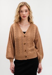 Blondynka w jasnobrązowym kardiganie z splotem w warkocz z guzikami i balonowymi rękawami, nosząca czarną bluzkę i spódnicę, stojąca na białym tle.