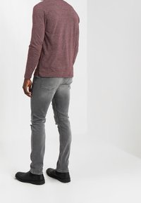 Camisa de manga larga en color burdeos, pantalones vaqueros gris desgastados y zapatos negros. La camisa presenta un escote redondeado y mangas ajustadas.