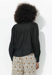 Blouse noire texturée avec des manches bouffantes et des poignets boutonnés, portée sur un pantalon crème à motifs avec des designs floraux et paisley.