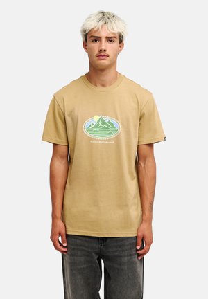 Jeune homme aux cheveux blonds clairs et moustache, portant un t-shirt beige avec un motif montagne et soleil et un jean gris foncé, debout sur fond blanc.