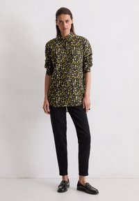 Chemise noire avec motif abstrait jaune et blanc, col �à boutons, manches longues, associée à un pantalon noir et des mocassins brillants.