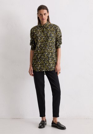 Chemise noire avec motif abstrait jaune et blanc, col à boutons, manches longues, associée à un pantalon noir et des mocassins brillants.