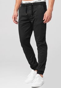 Black jogger pants made of lightweight cotton, featuring an elastic waistband with drawstring and elastic cuffs, paired with white sneakers.