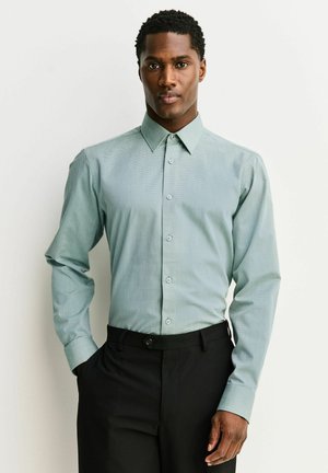 Homme portant une chemise à manches longues boutonnée vert clair à motifs, rentrée dans un pantalon noir habillé, debout avec une main dans la poche.