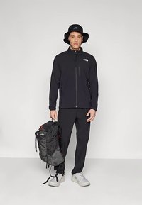 Svart glidelåsjakke med logo, matchende bukser, grå og svart ryggsekk, og svart bøttehatt. Modellen står mot en nøytral bakgrunn.