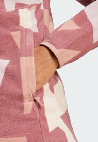 Prenda de forro polar con un patrón geométrico en tonos rosa y crema. Los detalles notables incluyen un bolsillo y una tela suave y texturizada.