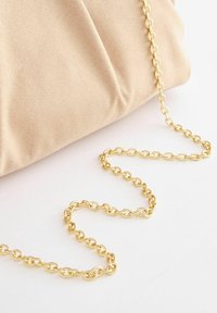 Beżowa torebka typu kopertówka o miękkiej fakturze, z złotym łańcuszkiem, który leży obok niej, ukazując okrągłe ogniwa.