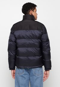 Pufferjack in marineblauw en zwart, met een hoge kraag, horizontale stiksels, elastische manchetten en een gladde textuur. Achteraanzicht dat het ontwerp toont.