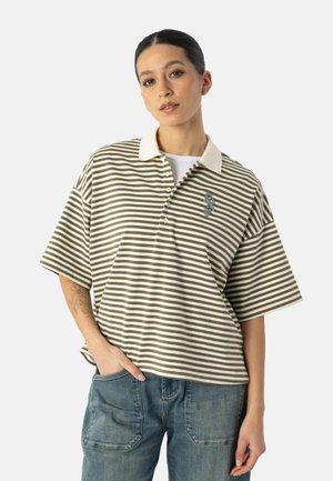 Frau mit zurückgebundenen Haaren trägt ein übergroßes olivgrün-weiß gestreiftes Poloshirt und blaue Denim-Jeans, Hand in der Tasche.