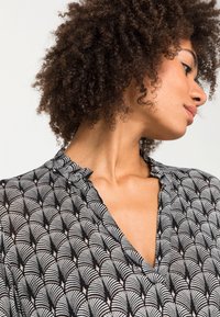 Blouse noire et blanche avec un motif en éventail, tissu doux, design en V, et détail de col froncé, avec des manches courtes.