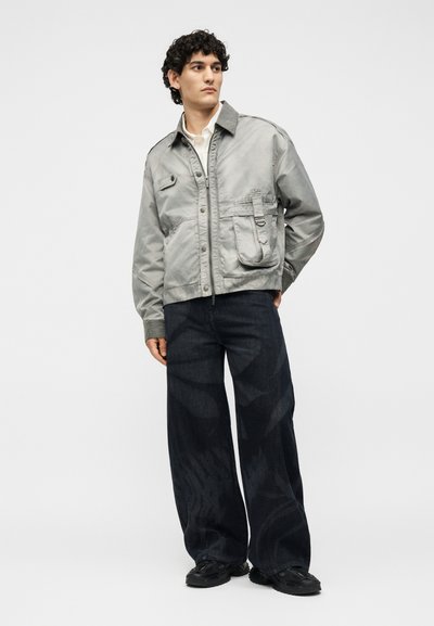 Νέος άνδρας που φοράει ανοιχτό γκρι εργοστασιακό τζάκετ, λευκό πουκάμισο, φαρδιά σκούρα τζιν και μαύρα παπούτσια, στέκεται με το ένα χέρι στην τσέπη.