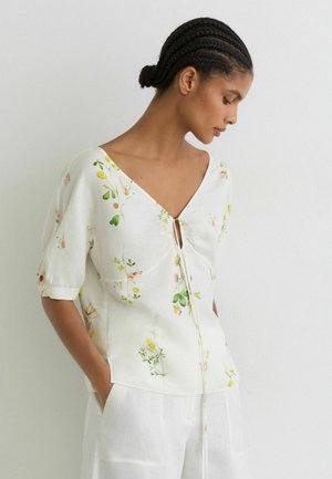Frau mit geflochtenem Haar, die ein weißes, florales V-Ausschnitt-Oberteil mit Bindendetail und weiße Hose trägt, steht vor einer einfarbigen hellen Wand.