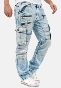 Pantalons cargo en denim bleu clair avec plusieurs poches zippées, des accents de côtes verticales et une coupe décontractée. Associés à des baskets blanches.