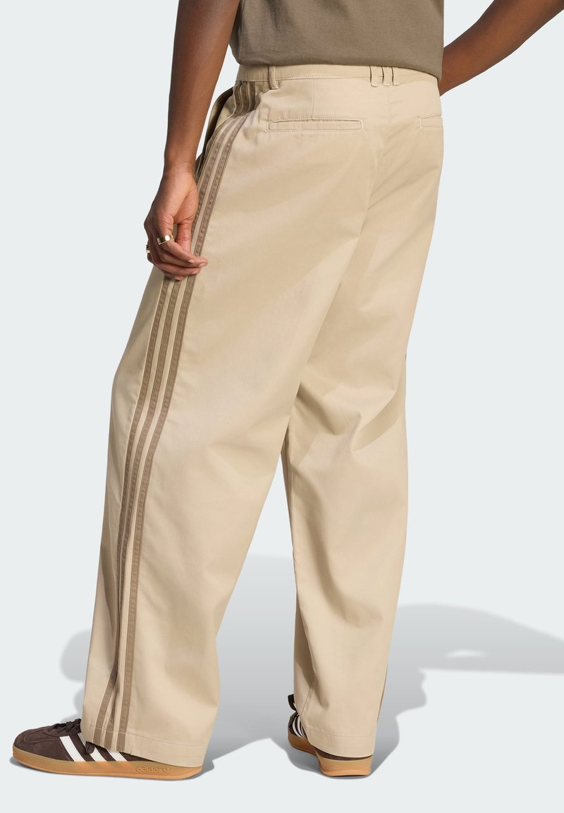 Person wearing beige wide-leg pants with brown side stripes and dark brown sneakers, standing facing away with hand near pocket.