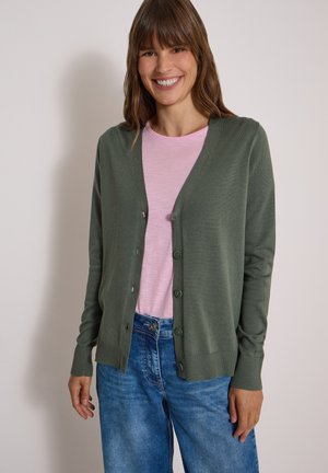 Femme souriante portant un cardigan vert boutonné par-dessus une chemise rose clair et un jean bleu, debout devant un fond clair uni.