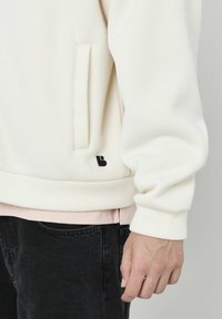 Sweatshirt côtelée couleur crème avec une poche latérale et un détail de logo noir. Le tissu semble doux, superposé sur une chemise rose, associé à un pantalon noir.