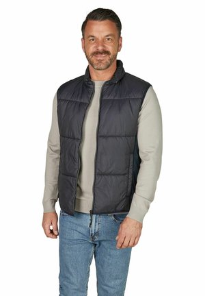 Smilende mand iført en sort quiltet vest over en beige sweater og blå jeans, stående mod en hvid baggrund.