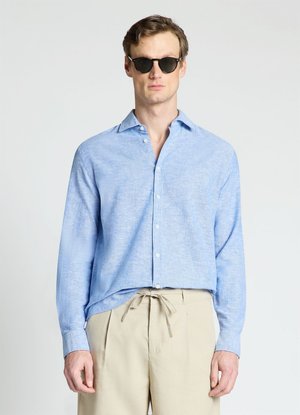 Uomo con occhiali da sole neri, camicia azzurro chiaro con bottoni e pantaloni beige con coulisse, in piedi davanti a uno sfondo semplice.