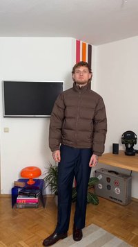 Giacca imbottita marrone con colletto alto, caratterizzata da una chiusura con zip e un design imbottito. Abbinata a pantaloni scuri e scarpe marroni.