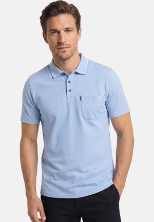 Mann trägt hellblaues kurzärmeliges Poloshirt mit Brusttasche und dunkler Hose, steht mit einer Hand in der Tasche vor weißem Hintergrund.