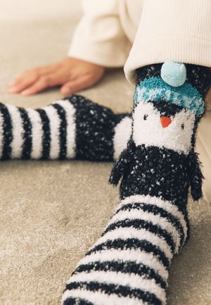 Picioare purtând șosete fuzzy alb-negru cu dungi și design cu pinguin pe un covor moale, o mână odihnindu-se în apropiere.