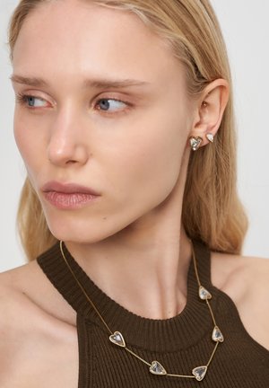 Goldene herzförmige Ohrringe und eine passende Halskette mit transparenten Herzakzenten, getragen mit einem dunkelgrünen, gerippten Oberteil.