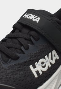 Μαύρο αθλητικό παπούτσι HOKA με υφασμάτινη υφή, μαύρα κορδόνια, λουράκι με βέλκρο και λευκό λογότυπο στο πλάι και στο λουράκι.