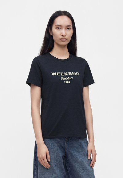 Femme aux longs cheveux noirs portant un t-shirt noir avec le texte "WEEKEND MaxMara 1983" et un jean bleu ample, debout devant un fond blanc uni.