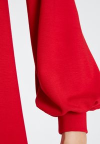 Roter Stoffärmel mit einem voluminösen, gerafften Design. Verfügt über einen taillierten Manschette am Handgelenk, glatte Textur und minimale Verzierungen.