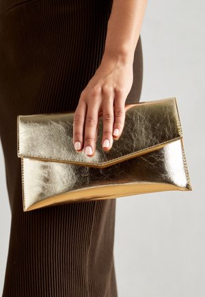 Gouden metallic clutch bag met een gladde textuur, envelopdesign en geplooide flapsluiting, vastgehouden in een hand tegen een neutrale achtergrond.