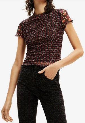 Woman wearing matching patterned black mesh top and pants with red and orange retro floral design, hand in pocket, against plain background.