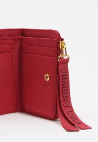 Raudonos medžiagos piniginės vidus su keliais kortelių skyreliais, auksinėmis detalėmis ir tekstūruota raudona juostele, ant kurios išrašytas tekstas "BIMBA Y LOLA."