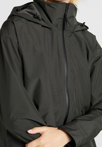 Vandtæt mørkegrøn jakke med høj krave, lynlåslukning, justerbar hætte og glat tekstur. Har minimal detaljering og en afslappet pasform.